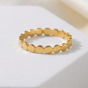 May include: A gold-colored ring featuring a series of small, circular, flat accents. The ring is set against a white background, with a soft, off-white fabric in the background. The ring's design is simple and elegant.