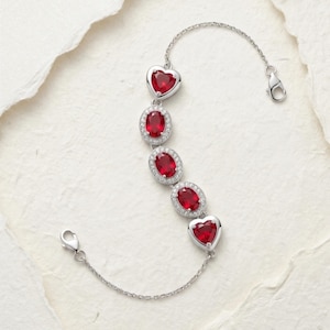 Pode incluir: Uma pulseira de tom prateado com cinco pedras preciosas vermelhas centrais, incluindo duas pedras em forma de coração nas extremidades e três pedras ovais no meio, cada uma rodeada por um halo de pequenas pedras claras e uma corrente fina.