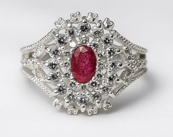 Puede incluir: Un anillo de plata con una gran piedra central de rubí ovalada, rodeada de piedras blancas más pequeñas. El anillo tiene un diseño intrincado y calado con piedras pequeñas adicionales a lo largo de la banda.