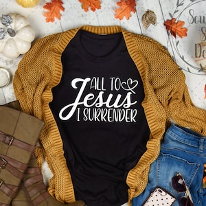 May include: A black t-shirt with the text "All to Jesus I Surrender" in white lettering. The shirt is styled with a brown cardigan, blue jeans, brown boots, and fall-themed decorations.