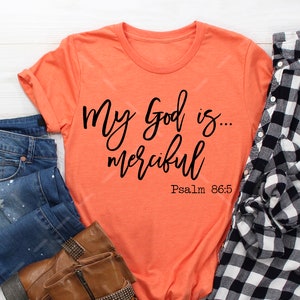 Könnte beinhalten: Orangefarbenes T-Shirt mit schwarzem Text, der "My God is... merciful" und "Psalm 86:5" lautet. Das Shirt liegt auf einer weißen Holzoberfläche mit blauen Jeans, braunen Stiefeln und einem schwarz-weiß karierten Hemd.