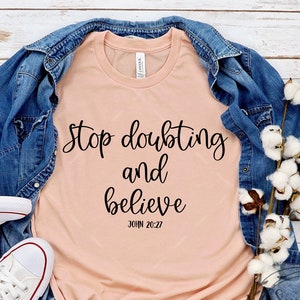 May include: A peach-colored t-shirt with the text "Stop doubting and believe" and "JOHN 20:27" in black lettering. The shirt is laid out on a white wooden surface with a denim jacket, white sneakers, and a sprig of cotton.