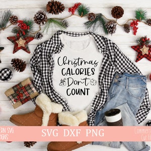 Puede incluir: Una camiseta blanca con texto negro que dice "Christmas Calories Don't Count". La camiseta está rodeada de tela a cuadros, jeans azules y decoraciones navideñas.