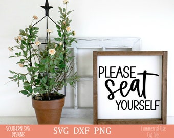 Puede incluir: Un letrero blanco con un marco de madera marrón, que muestra el texto "PLEASE seat YOURSELF" en fuente cursiva negra. El letrero se muestra en una superficie blanca con una planta en maceta en el fondo.