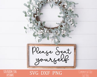 May include: A white wooden sign with the text "Please Seat Yourself" in black cursive font. The sign is sitting on a white wood background with a green and white wreath above it.