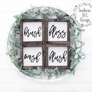 May include: Four wooden signs with white backgrounds and black calligraphy. The signs read "brush", "floss", "wash", and "flush". The signs are arranged in a square shape and are surrounded by a wreath of green leaves.