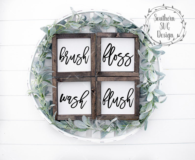 May include: Four wooden signs with white backgrounds and black calligraphy. The signs read "brush", "floss", "wash", and "flush". The signs are arranged in a square shape and are surrounded by a wreath of green leaves.
