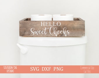 May include: A wooden box with the text "Hello Sweet Cheeks" on it. The box is sitting on top of a white toilet tank and is holding two rolls of toilet paper.