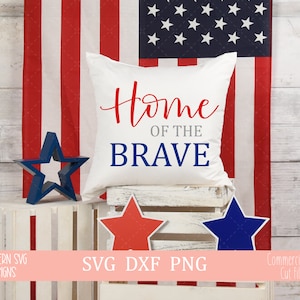 Puede incluir: Una almohada blanca con el texto "Home of the Brave" en letras rojas, azules y grises. La almohada está sentada en una caja de madera blanca con una estrella roja y azul a cada lado. Una bandera estadounidense está en el fondo.