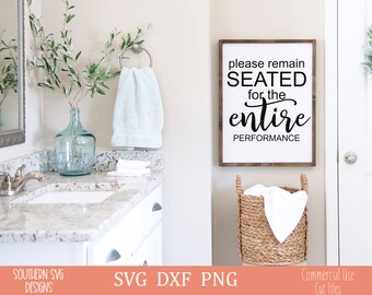 May include: A white bathroom sign with black text that reads "Please remain seated for the entire performance". The sign is hanging on a white wall in a bathroom with a white vanity and a blue glass vase with greenery.