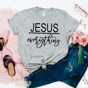 May include: A gray t-shirt with the text "JESUS everything" in black lettering. The shirt is styled with blue jeans, pink shoes, and a bouquet of pink flowers.