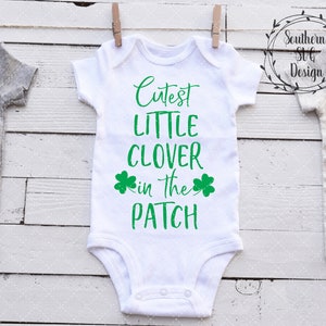 May include: White baby onesie with green shamrock designs and the text "Cutest Little Clover in the Patch".