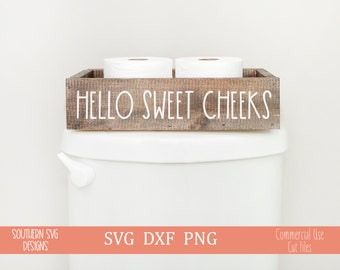 May include: A wooden box with the text "Hello Sweet Cheeks" on the front. The box is sitting on top of a white toilet tank and contains two rolls of toilet paper.
