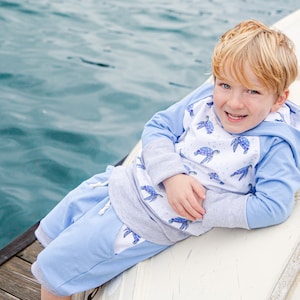 May include: A young boy wearing a light blue hooded sweatshirt with a white and blue turtle print, and matching light blue shorts, is sitting on a white boat. He is smiling and looking at the camera.