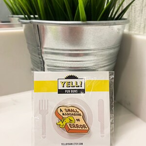 May include: A yellow and brown enamel pin with a slice of bread with a pat of butter on it. The text on the pin reads "A Small Margarine of Error". The pin is packaged on a white card with the text "Yell! Pun Buns" and a website address.