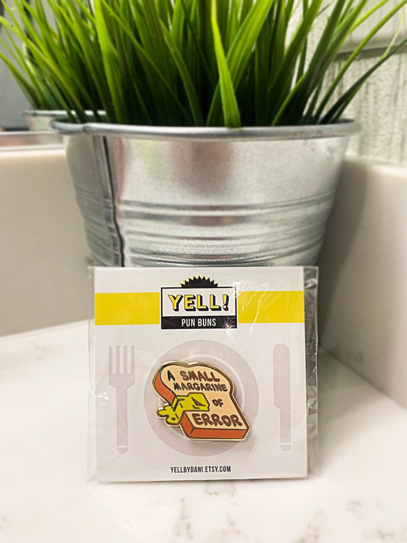 May include: A yellow and brown enamel pin with a slice of bread with a pat of butter on it. The text on the pin reads "A Small Margarine of Error". The pin is packaged on a white card with the text "Yell! Pun Buns" and a website address.
