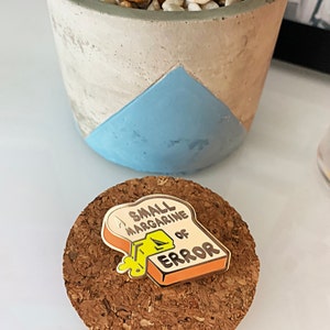 May include: A small enamel pin shaped like a slice of bread with the text "A Small Margarine of Error" on it. The pin is yellow, orange, and white.