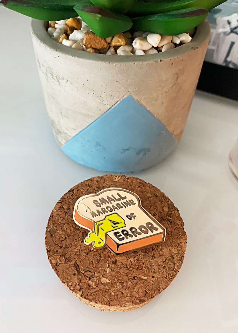 May include: A small enamel pin shaped like a slice of bread with the text "A Small Margarine of Error" on it. The pin is yellow, orange, and white.