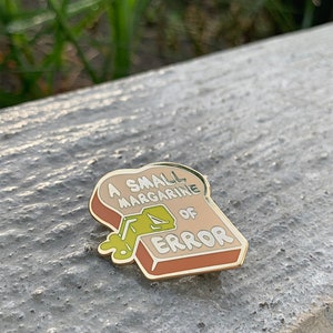 May include: A gold-colored enamel pin shaped like a slice of bread with the text "A small margarine of error" and a green cartoon of a stick of margarine spilling out of the bread.