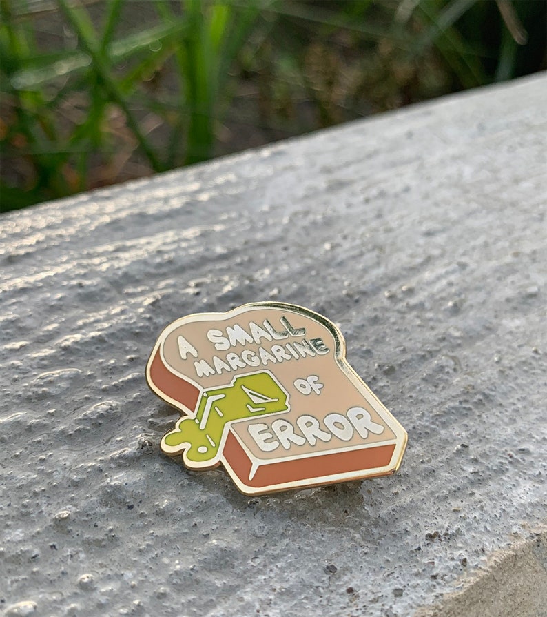 May include: A gold-colored enamel pin shaped like a slice of bread with the text "A small margarine of error" and a green cartoon of a stick of margarine spilling out of the bread.