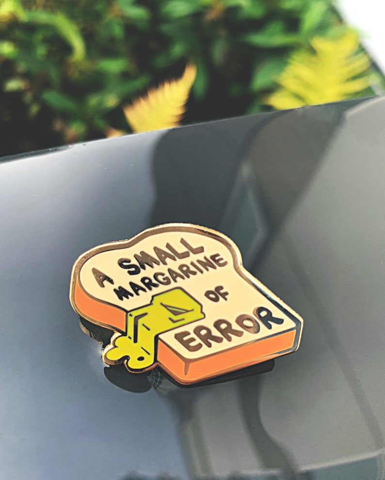 May include: A gold-colored enamel pin shaped like a slice of bread with the text "A small margarine of error" and a yellow stick of margarine melting on top.