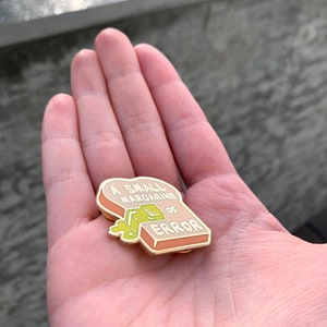 May include: A gold-colored enamel pin shaped like a slice of bread with the text "A small margarine of error" and a green blob of margarine dripping from the top.