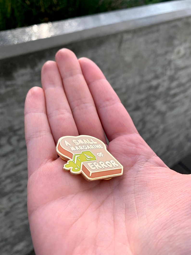 May include: A gold-colored enamel pin shaped like a slice of bread with the text "A small margarine of error" and a green blob of margarine dripping from the top.
