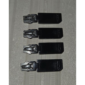 May include: Four black zipper pulls with silver-colored metal zipper heads. The zipper pulls are rectangular and have a small, angled design on the end. The background is a light gray and white striped fabric.