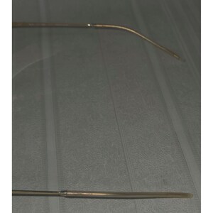 Puede incluir: Un par de monturas de gafas doradas. Las monturas tienen una forma rectangular delgada con una patilla curva. Las gafas est&aacute;n sobre un fondo gris.