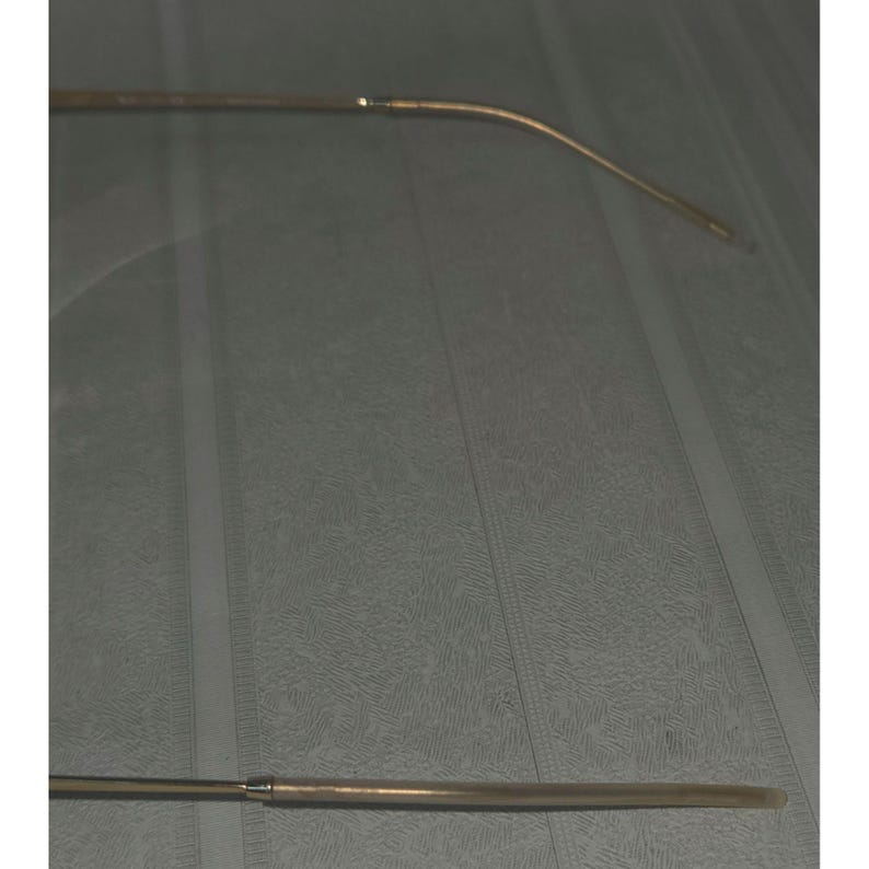 Puede incluir: Un par de monturas de gafas doradas. Las monturas tienen una forma rectangular delgada con una patilla curva. Las gafas est&aacute;n sobre un fondo gris.