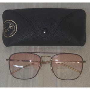 Puede incluir: Gafas con montura dorada y lentes de color rosa, con un estuche de cuero negro. Las gafas tienen doble puente y un estilo aviador cl&aacute;sico. El estuche tiene un cierre de bot&oacute;n y un logotipo dorado.