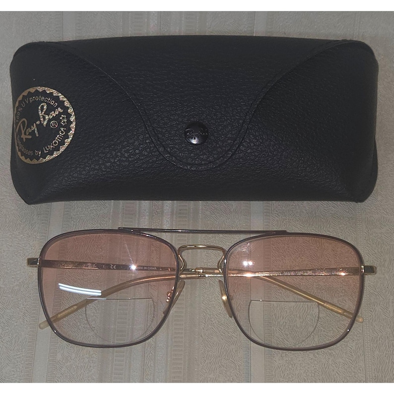 Puede incluir: Gafas con montura dorada y lentes de color rosa, con un estuche de cuero negro. Las gafas tienen doble puente y un estilo aviador cl&aacute;sico. El estuche tiene un cierre de bot&oacute;n y un logotipo dorado.