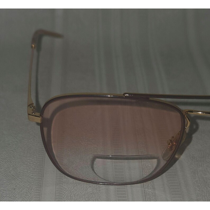 Puede incluir: Gafas de sol con montura dorada y lentes tintadas en marr&oacute;n. Las gafas tienen una forma de aviador cl&aacute;sica con doble puente y barra superior curvada. Las lentes tienen un color degradado, que pasa de un tono m&aacute;s oscuro en la parte superior a uno m&aacute;s claro en la inferior.
