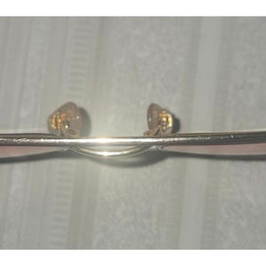 Puede incluir: Primer plano de gafas doradas con puente curvo y lentes rosas. Las almohadillas nasales son de color marr&oacute;n claro. Las gafas est&aacute;n sobre un fondo blanco.