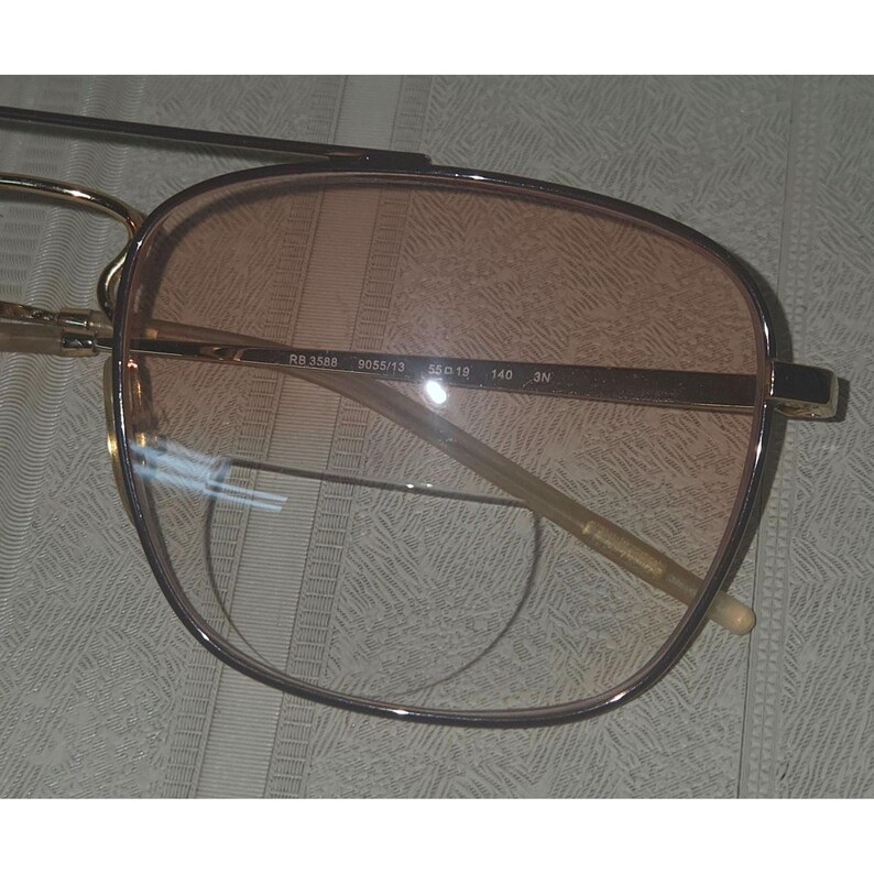 Puede incluir: Gafas de sol con montura dorada y lentes degradadas. Las lentes hacen una transici&oacute;n de un melocot&oacute;n claro a un tono m&aacute;s oscuro. La montura tiene forma rectangular con un dise&ntilde;o de doble puente. Las patillas son finas y doradas, con texto impreso en la montura.
