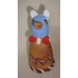 May include: A ceramic figurine of a bird with a blue head, white ears, and a red bow tie. The bird is standing on a brown base with white and blue painted designs.