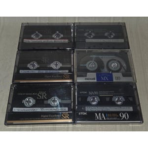 May include: A collection of six vintage audio cassette tapes in clear plastic cases. The tapes feature labels with text such as "SR", "MA90", and "MX 110". The tapes are black and silver, with various text and logos.