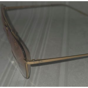 Puede incluir: Gafas de sol con montura dorada y lentes marrones claros. El logotipo de Ray-Ban es visible en la patilla. Las gafas tienen forma rectangular.