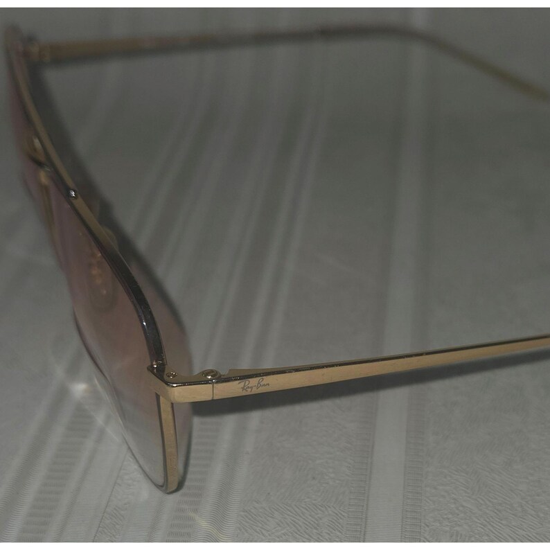 Puede incluir: Gafas de sol con montura dorada y lentes marrones claros. El logotipo de Ray-Ban es visible en la patilla. Las gafas tienen forma rectangular.