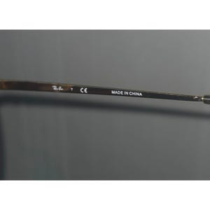 Puede incluir: Primer plano de una patilla de gafas de sol con el texto "MADE IN CHINA". La patilla es de color met&aacute;lico oscuro con el logotipo de la marca y la marca "CE". El fondo es gris liso.