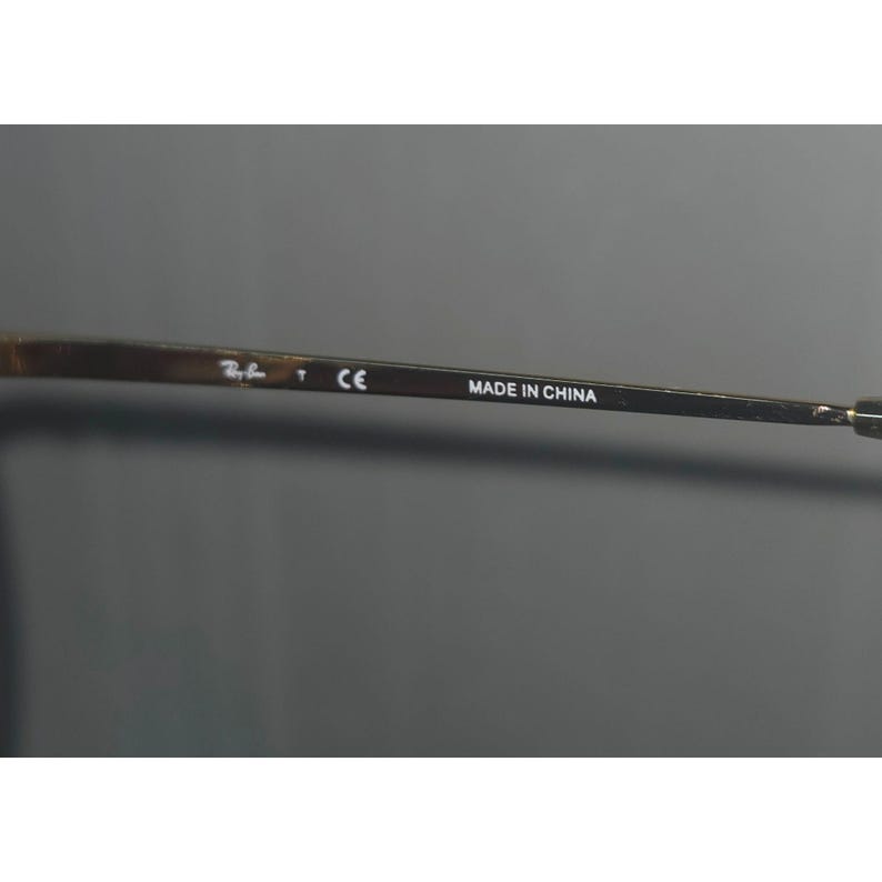 Puede incluir: Primer plano de una patilla de gafas de sol con el texto "MADE IN CHINA". La patilla es de color met&aacute;lico oscuro con el logotipo de la marca y la marca "CE". El fondo es gris liso.