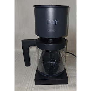 May include: A dark grey coffee maker with a clear glass carafe. The digital display shows "12:00 AM". The handle is on the left side. The base is square. The words "Brew | Stop" are below the time.
