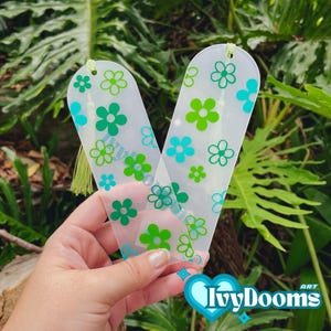 Op de afbeelding: Twee transparante plastic bladwijzers met een bloemenmotief. De bladwijzers hebben groene en blauwe bloemen op een witte achtergrond. De bladwijzers zijn doorschijnend en hebben een kwastje aan de bovenkant.