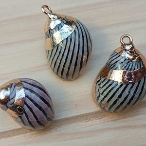 Peut inclure: Trois coquillages à rayures noires et blanches ornés de dorure. Les coquillages sont enfilés sur une chaîne dorée et sont prêts à être utilisés comme pendentifs.