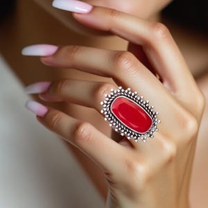 Op de afbeelding: Een zilveren ring met een grote, ovale rode edelsteen gezet in een decoratief, versierd frame.