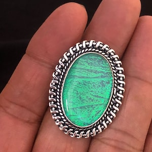 Puede incluir: Anillo ovalado con una piedra verde brillante y un diseño texturizado. La piedra está engastada en un marco de metal plateado con un borde detallado en forma de cuerda. Una joya.