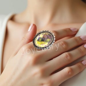 Op de afbeelding: Een ring met een grote, ovale, gefacetteerde iriserende steen met gele en paarse tinten, gezet in een zilverkleurige metalen band met een gedraaide rand. Het woord Joyery is in witte letters zichtbaar in het midden van de ring.