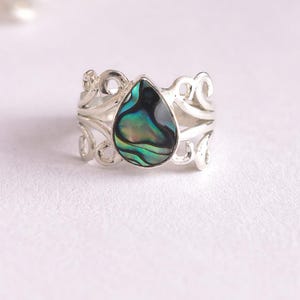 May include: A silver ring with an abalone teardrop-shaped stone. The ring has an ornate, openwork design around the band. The abalone stone displays iridescent blues, greens, and blacks. The ring is set against a white background.