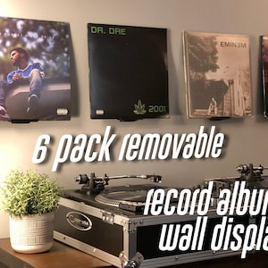 May include: Six black wall mounts for displaying vinyl records. The mounts are attached to a wall with four records displayed. The records are "2014 Forest Hills Drive" by J. Cole, "2001" by Dr. Dre, "Eminem" by Eminem, and an unknown record. A turntable is in the foreground.