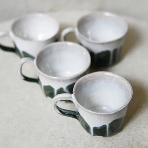 May include: A set of four ceramic mugs featuring a white glazed interior and upper exterior, transitioning into a dark green drip glaze pattern on the lower half. Each mug has a single curved handle and a slightly irregular, organic rim shape.